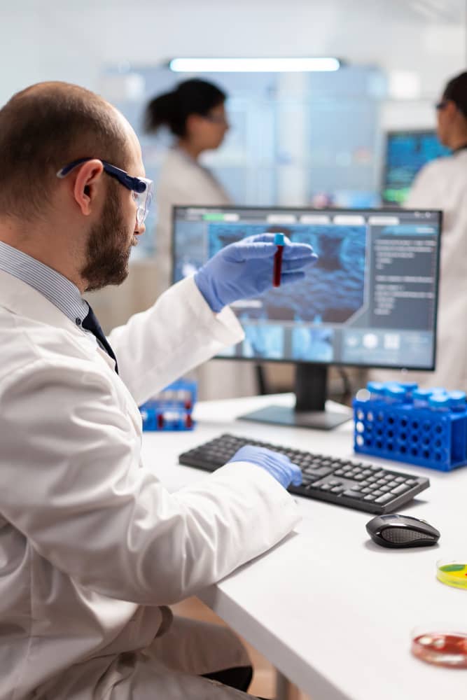 A bald man with a beard wearing glasses, blue latex gloves, and a white lab coat examines a test tube with a red substance in a scientific laboratory. In the background, screens display molecular structures and a female colleague works beside another male colleague at Healthcare Industry Page image in the Tanyo CRM webiste.