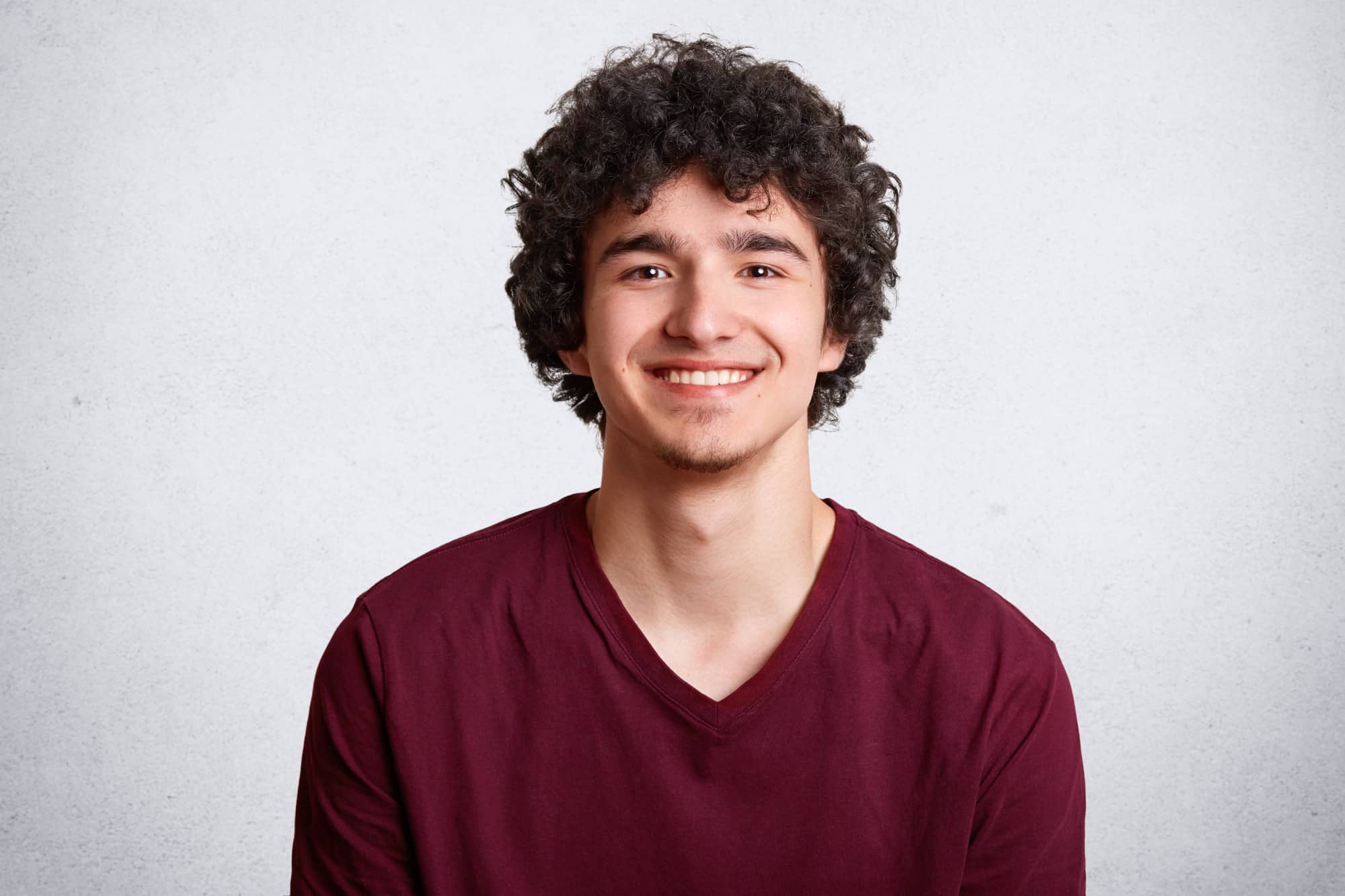 Young man with curly dark hair smiling, wearing a maroon v-neck t-shirt, against a textured light gray background define feature customer cofounder in the Tanyo CRM webiste.