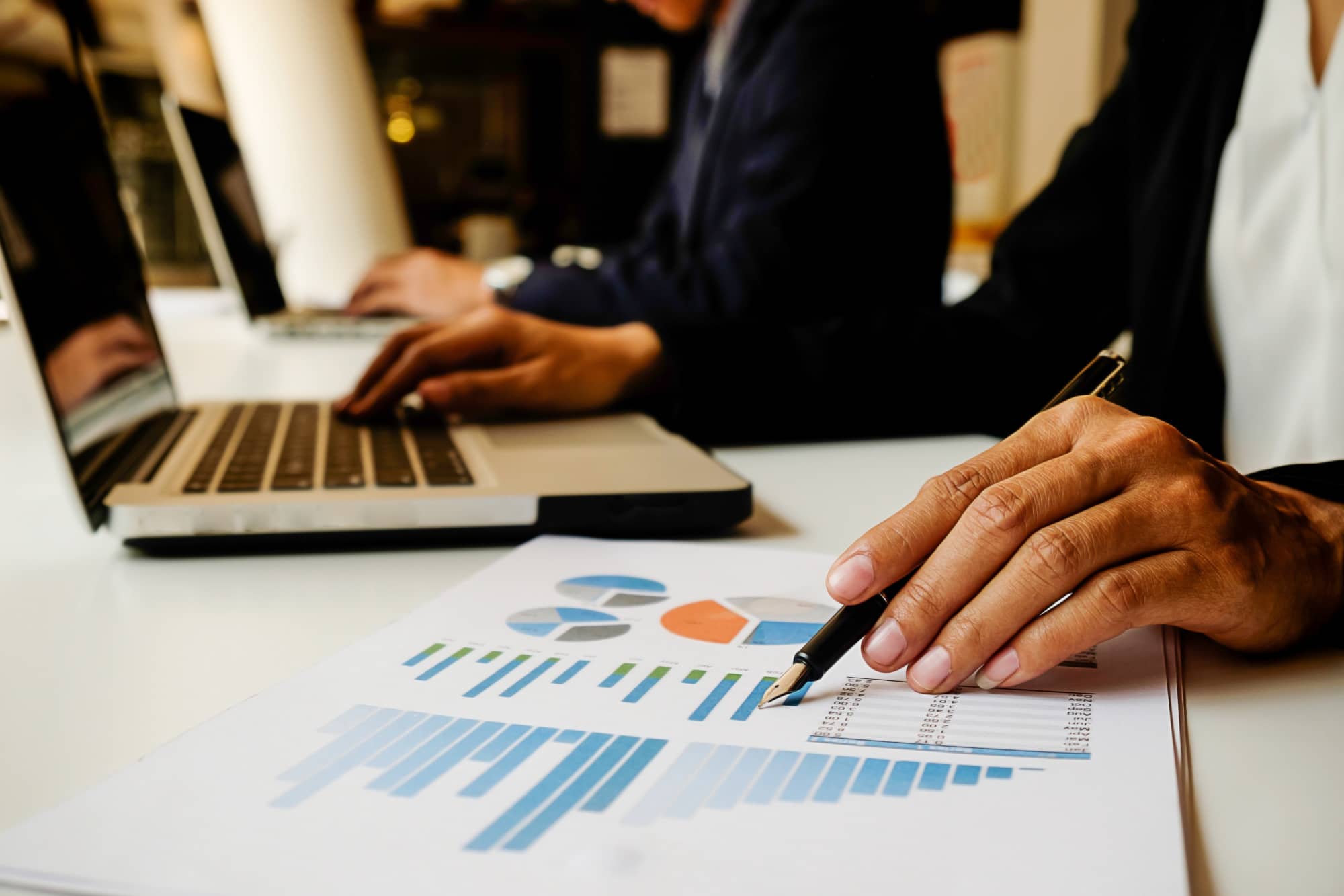 Close-up of a person's hand holding a pen over a printed chart with various graphs, with another individual typing on a laptop in the background. define finance consulting discussing executive people plan image in the Tanyo CRM webiste.