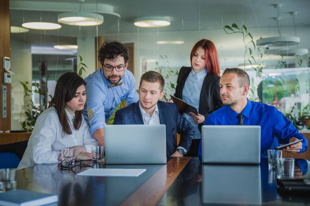 Five professionals collaborating around a conference table in a modern office setting, using laptops and focusing intently on their work, with green plants in the background industry page background mobile image in the Tanyo CRM webiste.