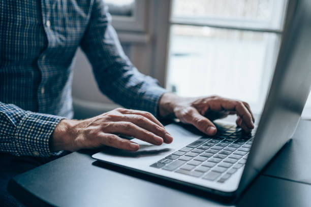 Close-up of an older man's hands typing on a laptop keyboard, visible in a plaid shirt, sitting at a desk by a window image in the Tanyo CRM webiste.